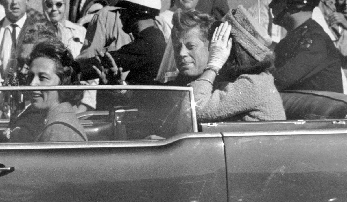 Lámhachadh JFK marbh in Dallas ar an 22 Samhain 1963 os comhair fhoirgneamh Texas School Book Depository